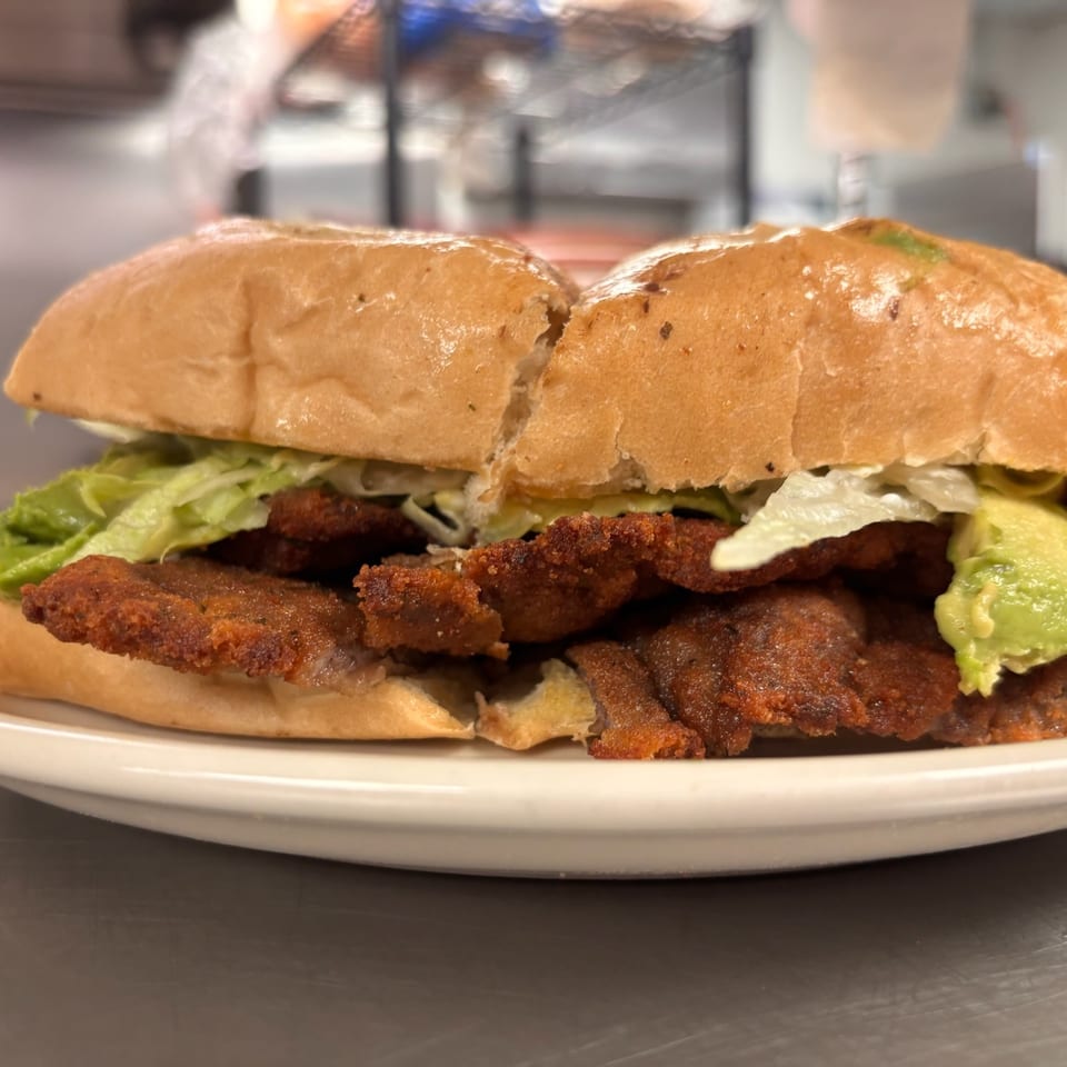Close-up of a toasted sandwich filled with crispy fried meat, lettuce, avocado, and sauce on a white plate.