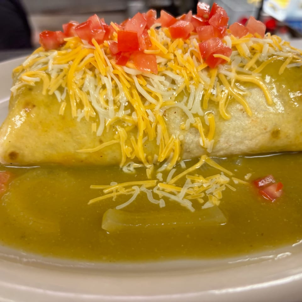 Large burrito smothered in green sauce, topped with shredded cheese and diced tomatoes on a white plate.