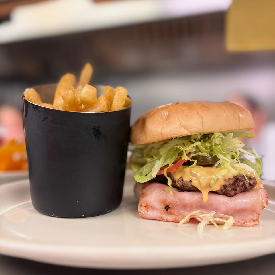 Cheeseburger topped with lettuce, tomato, sauce, and bacon served beside golden fries in a black cup.