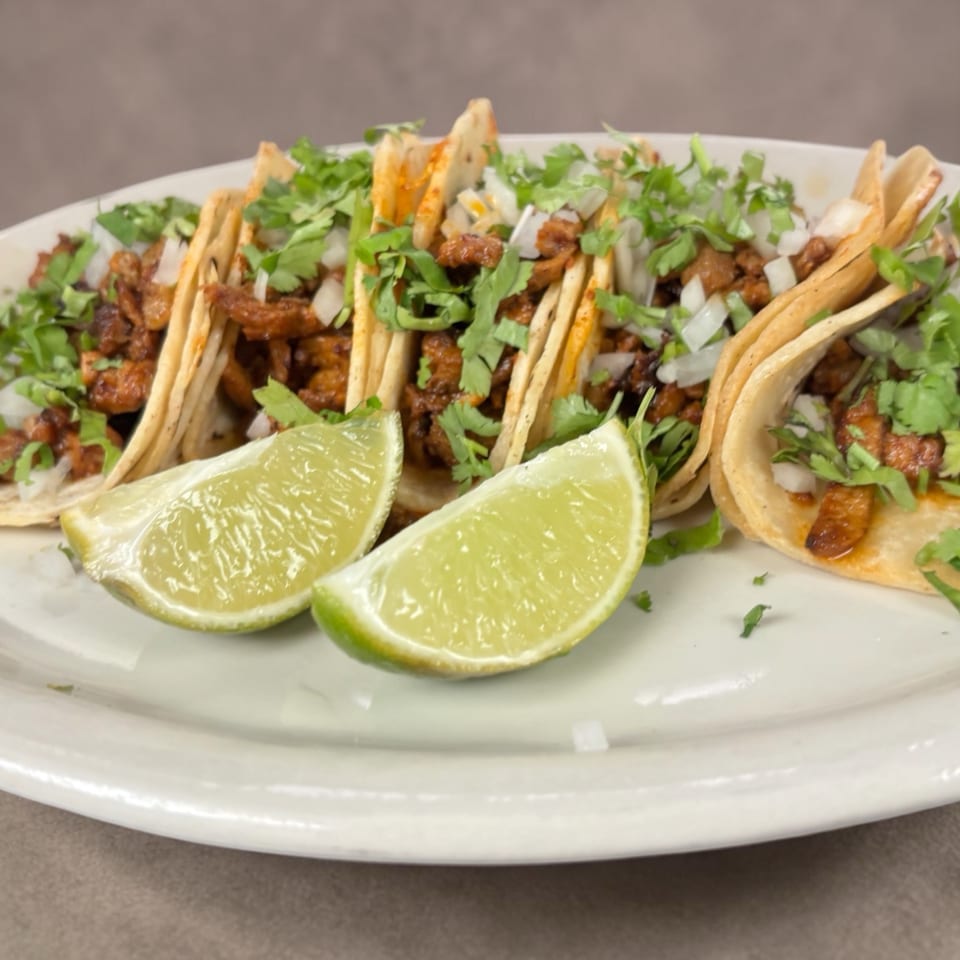 Plate of crispy tacos filled with seasoned meat, topped with cilantro and onions, with lime wedges in front.