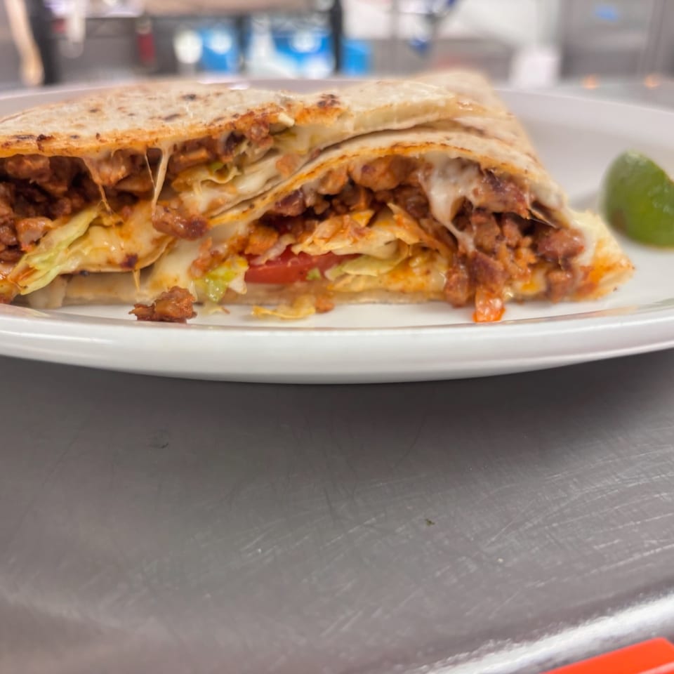 Grilled folded quesadilla filled with seasoned meat, melted cheese, lettuce, and tomato, served with a lime wedge.